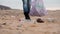 Volunteer walks on a sandy beach and collects garbage in a plastic bag. Legs close-up. Low angle. The concept of