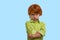 Waist up emotional portrait of redhead boy wearing green shirt on a blue background in the studio. Hi standing arms crossed and