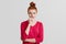 Waist up portrait of serious pretty female model with ginger hair knot wears red sweater, keeps hand under chin, listens seriously