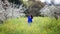 Waist up portrait of young woman in bright blue top in blossoming orchard and yellow mustard field, smiling, looking down off the