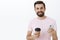 Waist-up shot of amused and happy delighted good-looking man having break holding smartphone and paper cup of coffee