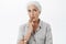 Waist-up shot of creative and thoughtful charming elderly mother with white hair biting finger and gazing focused at