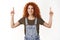 Waist-up shot friendly ambitious redhead curly woman show upper promo, pointing up to top, smiling delighted, introduce