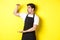 Waiter in black apron looking amazed at something large, holding big object, standing over yellow background