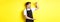 Waiter in black apron looking amazed at something large, holding big object, standing over yellow background