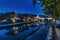 Walkway alongside a calm river with reflections of trees, light, and houses on the water