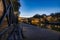 Walkway alongside a calm river with reflections of trees, light, and houses on the water