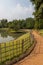 Walkway in a park with fence and pond in one side and trees and siting bench on the other