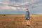 A wandering guy in a hoodie in a hood looks thoughtfully at a wheat field with a backdrop of the setting sun of a stormy sky