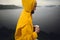 Wanderlust. Traveler in yellow raincoat holding metal mug and standing on cliff  in rainy windy day with view on lake. Hipster man