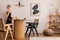 Warm and cozy interior of living room space with mock up poster frame, round table, rattan chairs, pedant lamp and walking young