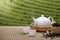 Warm cup of tea with teapot, green tea leaves and dried herbs on the bamboo mat at morning in plantations background with empty