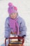 Warmly dressed smiling girl in pink scarf and hat relies on sled