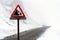 Warning sign red triangle falling rocks on icy snow covered winding mountain road passing through foggy and mist filled valley