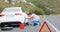 Warning triangle in rural road with man kneeling by broken-down white car defocussed in background