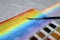 A watercolor brush is beyond a rainbow and rainbow painting on a watercolor block. A set of used solid watercolor paints is beside