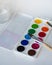 Watercolors of twelve colors in a box and brushes for painting, as well as a jar of water for painting on a white sheet