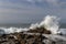 Wave crashing on rocks in foreground; ocean, cloudy blue sky in background.