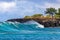 Wave on Hawaiian coast; trees on rocky shore. Clouds in background.