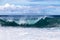 Wave on Hawaiian coast; trees on rocky shore. Clouds in background.