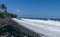 Waves attack the stony beach, and sea water spills over the beach`s edge. Palm trees and tropical vegetation line the coast of the