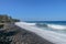 Waves attack the stony beach, and sea water spills over the beach`s edge. Palm trees and tropical vegetation line the coast