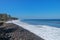 Waves attack the stony beach, and sea water spills over the beach`s edge. Palm trees and tropical vegetation line the coast