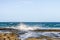 Waves crashing against flat lava rocks jutting into the ocean with freight ships dotting the horizon