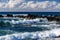 Waves on Hawiian shore; volcanic rocks in surf. Pacific in distance, with blue sky and clouds.