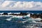 Waves on Hawiian shore; volcanic rocks in surf. Pacific in distance, with blue sky and clouds.