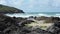 Waves rolling into a rocky cove, with coastal path in distance