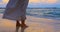 Waves washing over feet and wind blows woman dress who is standing on shore at epic sunset. Calm and chill atmosphere is