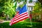 Waving American Flag large  in front of blurred upscale two story landscaped home with car behind security gate surrounded by