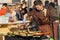 Weary cook prepares meat dishes in cast iron pans in outdoor kitchen