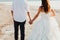 Wedding couple is holding hands and walking away on the sea beach. Sunny summer photo. Bride with hair down in off shoulder dress