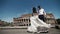 Wedding couple in Rome dancing barefoot at the rooftop near Coliseum, happy groom watches his bride playing with her