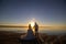 Wedding couple on the sea beach watching sunset. Sunny summer photo. Bride with hair down in off shoulder dress with train. Ocean
