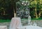 Wedding glass arch with white patterns decorations is standing outdoors in the green summer park. Pink hortensia and roses flowers