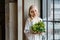 Wedding morning of the bride. Exquisite blonde in a delicate peignoir standing near the window with a bouquet of flowers in her