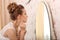 Wedding preparation. Beautiful young bride in white wedding dress indoors. Luxuty model looking at mirror and put on earring, at h