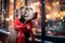 A Weimaraner dog wearing a red Christmas scarf against the backdrop of a city street, next to a Christmas store window