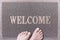 Welcome Door Mat With Female Feet. Friendly Grey Door Mat Closeup with Bare Woman Feet Standing. Welcome Carpet.