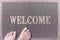 Welcome Door Mat With Female Feet. Friendly Grey Door Mat Closeup with Bare Woman Feet Standing. Welcome Carpet.