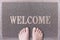 Welcome Door Mat With Female Feet. Friendly Grey Door Mat Closeup with Bare Woman Feet Standing. Welcome Carpet.