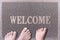 Welcome Door Mat With Three Feet. Friendly Grey Door Mat Closeup with Bare Feet Standing. Welcome Carpet.