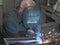 Welder in work clothes, construction gloves and a welding mask is welded with a welding machine metal product table,