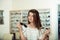 Which one is better fit me. Portrait of cheerful hesitating woman in optician store, making decision, holding stylish