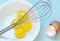 The whisk over fresh raw chicken yolks, eggs shells on the blue background. Baking or food preparation concept