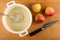 Whisk in white pan with dough, apples, kitchen knife on wooden table. Top view