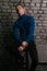 white attractive guy sits on a brick wall background dressed in a blue sweater and black pants